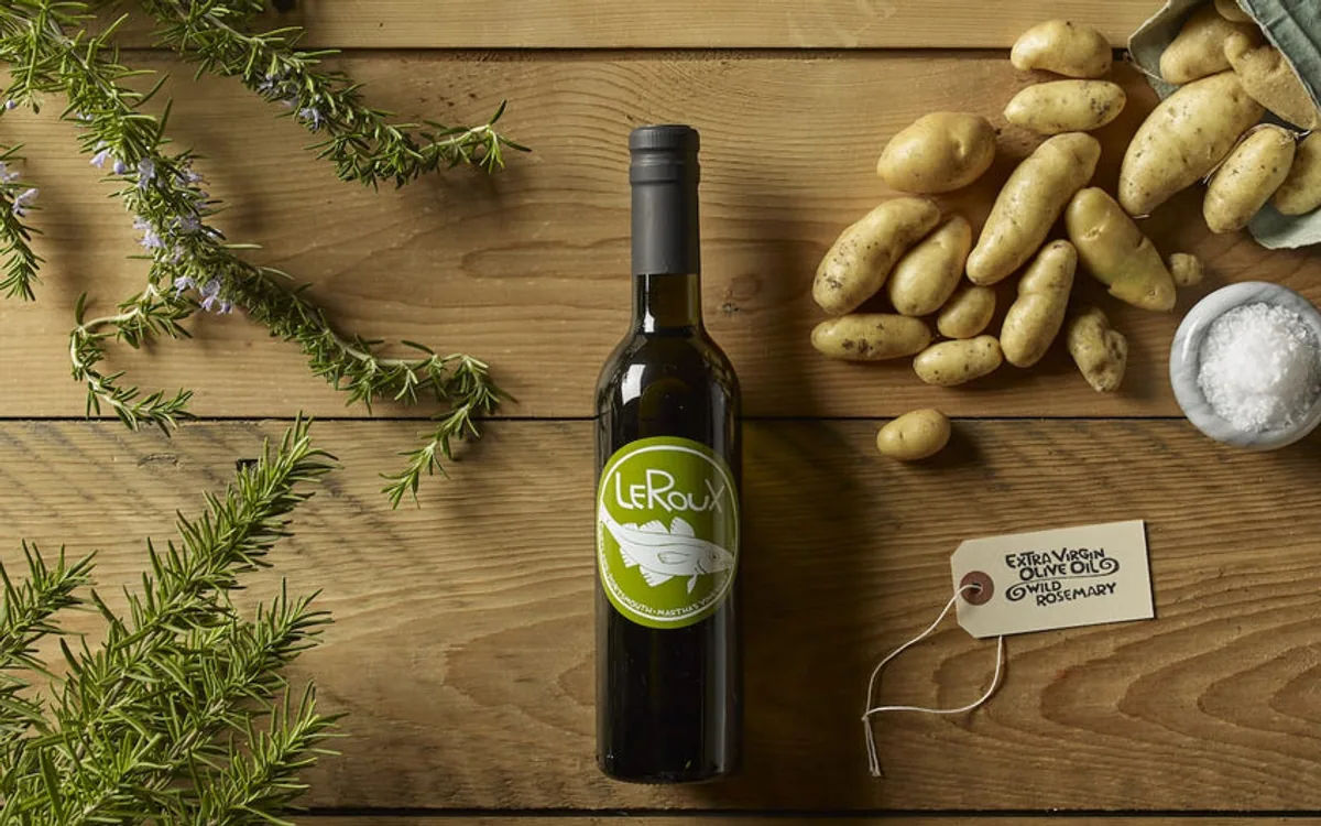 Extra virgin olive oil bottle with rosemary sprigs, potatoes, and salt on wooden table.
