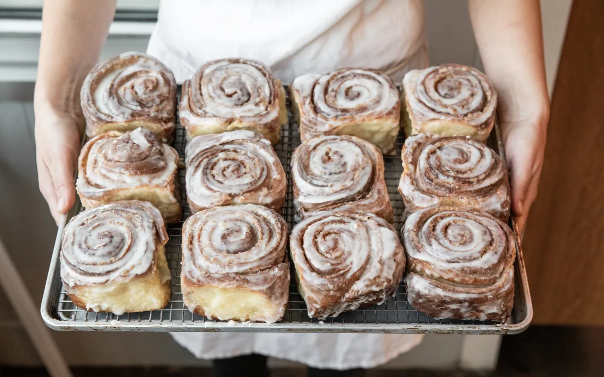 A person in a white apron holding a tray of freshly baked cinnamon rolls, each with a swirl pattern and glazed topping.